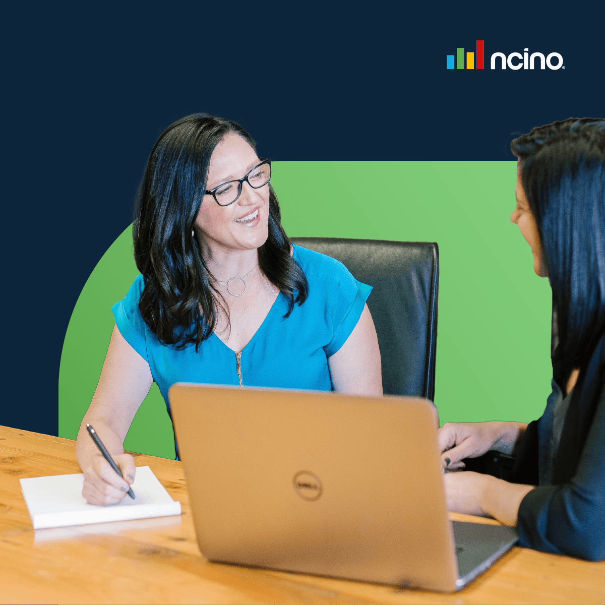 A woman in a blue shirt smiles at another woman over a laptop.