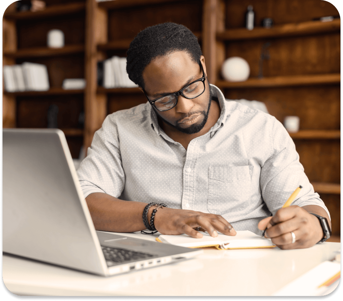 A man writes on a piece of paper with his laptop open on the desk.