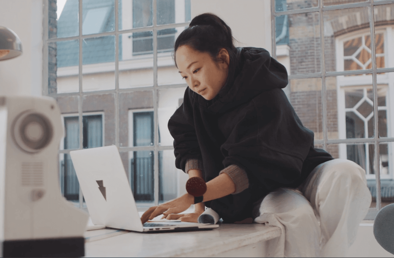 A wants in a dark hoodie sits at a table as she works on her laptop in her studio. 