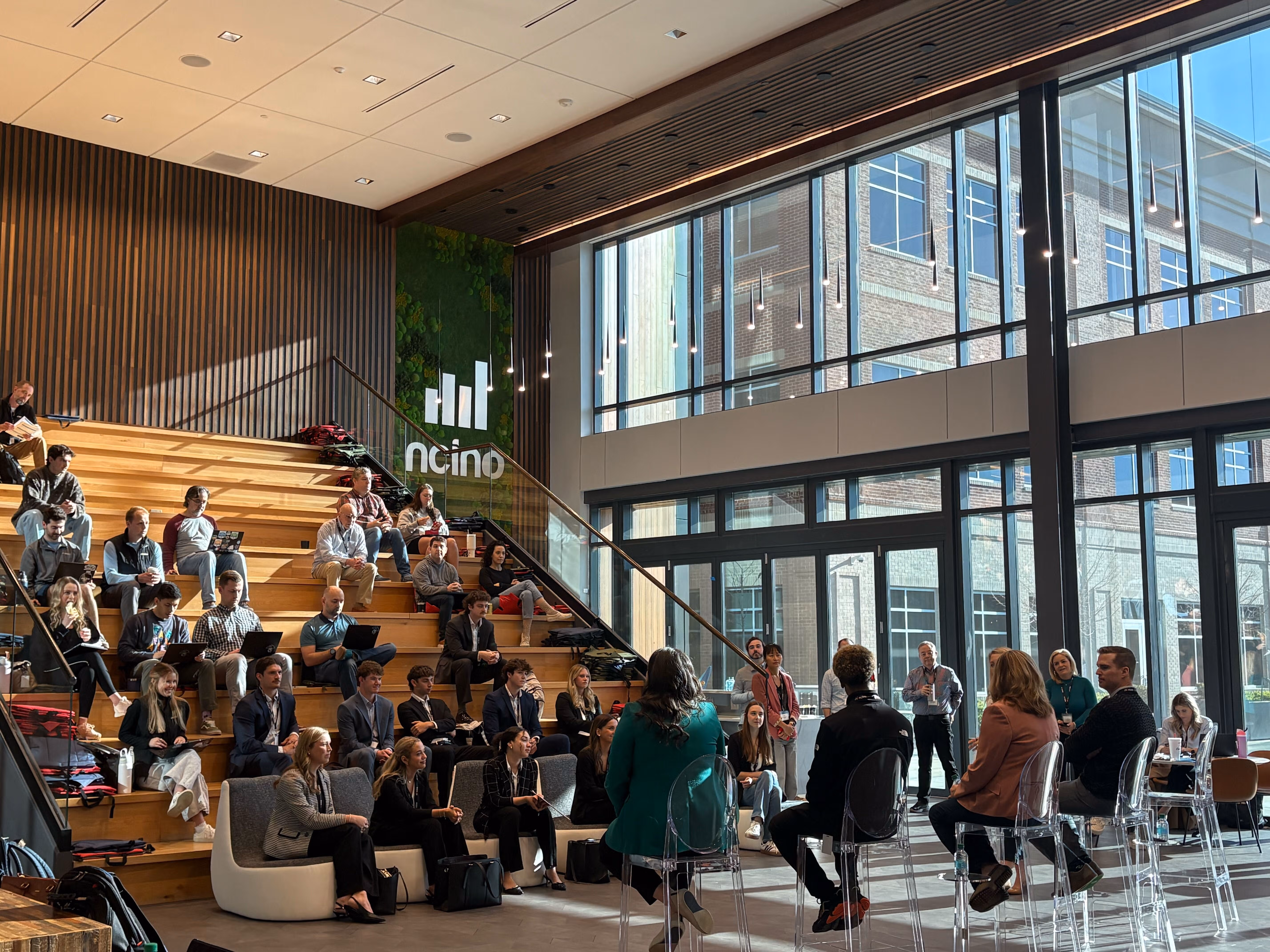 A crowd of students, faculty, and nCino employees at nCino headquarters watching the final round of the UNCW x nCino Sales Showdown, with a student presenting to a panel of judges seated in the foreground.