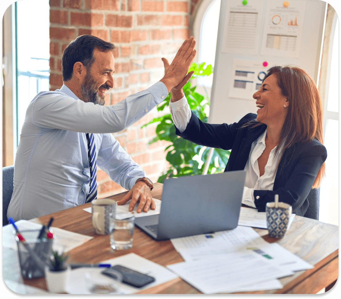 Two coworkers exchange a celebratory high-five in front of a laptop and a bunch of papers.